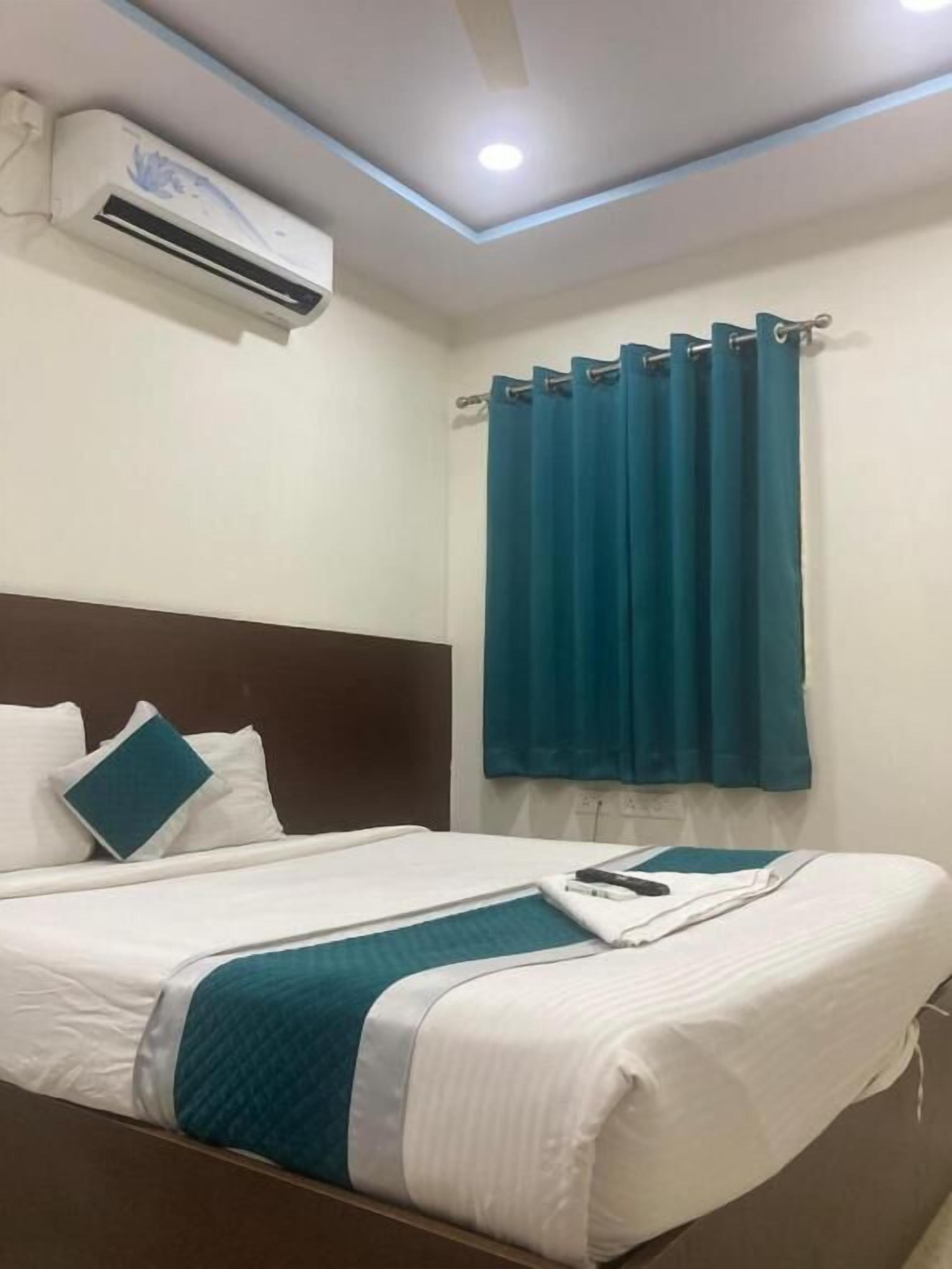 A hotel room with a bed, white bedding, a teal throw, a brown headboard, a window with teal curtains, and an air conditioning unit.