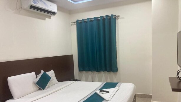 Een hotelkamer met een bed, wit beddengoed, een blauwgroene plaid, een gordijn en een airconditioner.