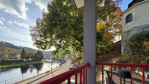 Ein Balkon mit roter Brüstung, der auf einen Fluss, ein Steinhaus und Herbstlaub blickt.