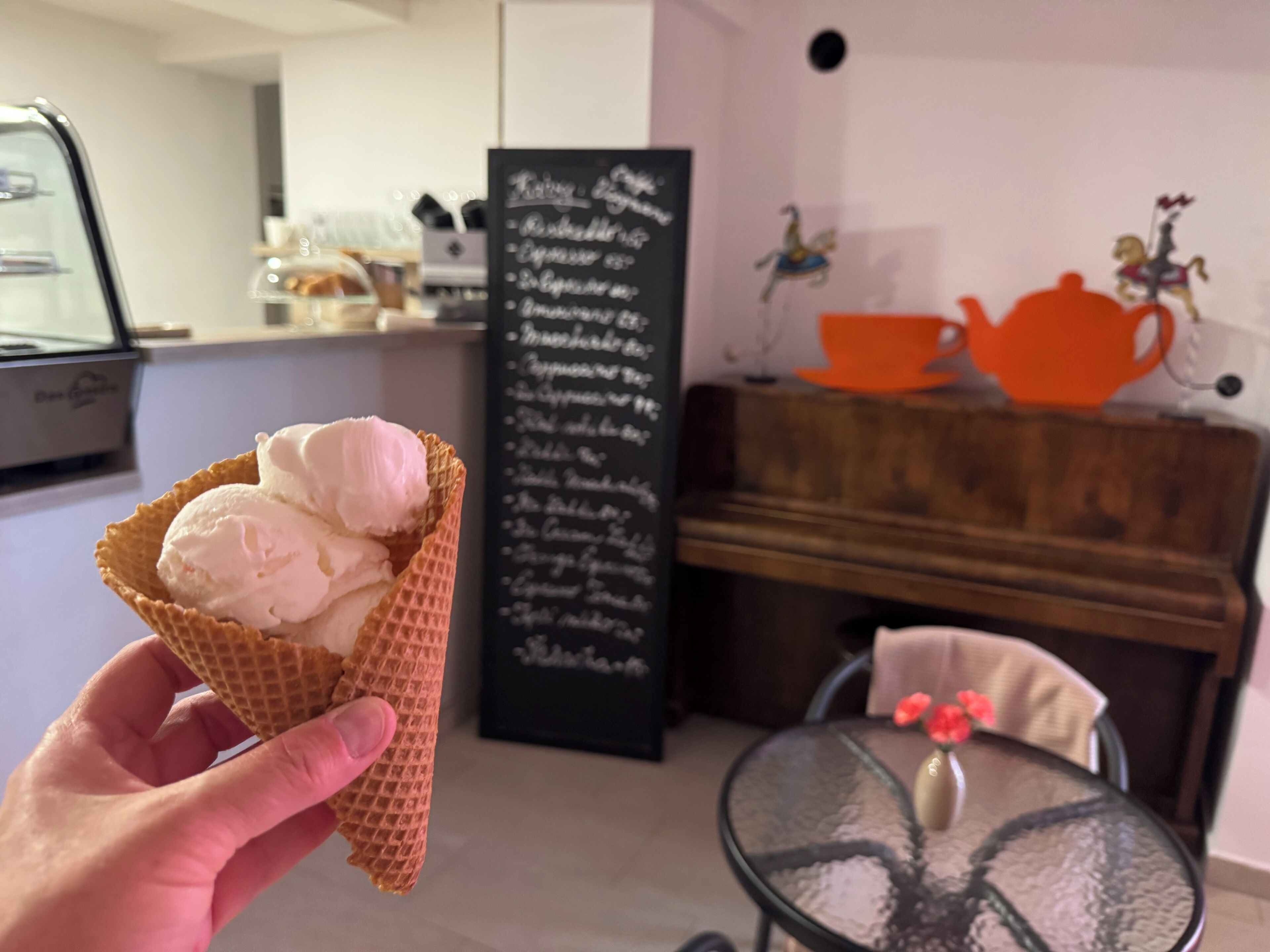 Une main tenant un cornet de gaufre garni de trois boules de glace. En arrière-plan, on aperçoit un tableau noir servant de menu, un piano en bois et une table sur laquelle est posé un vase de fleurs.