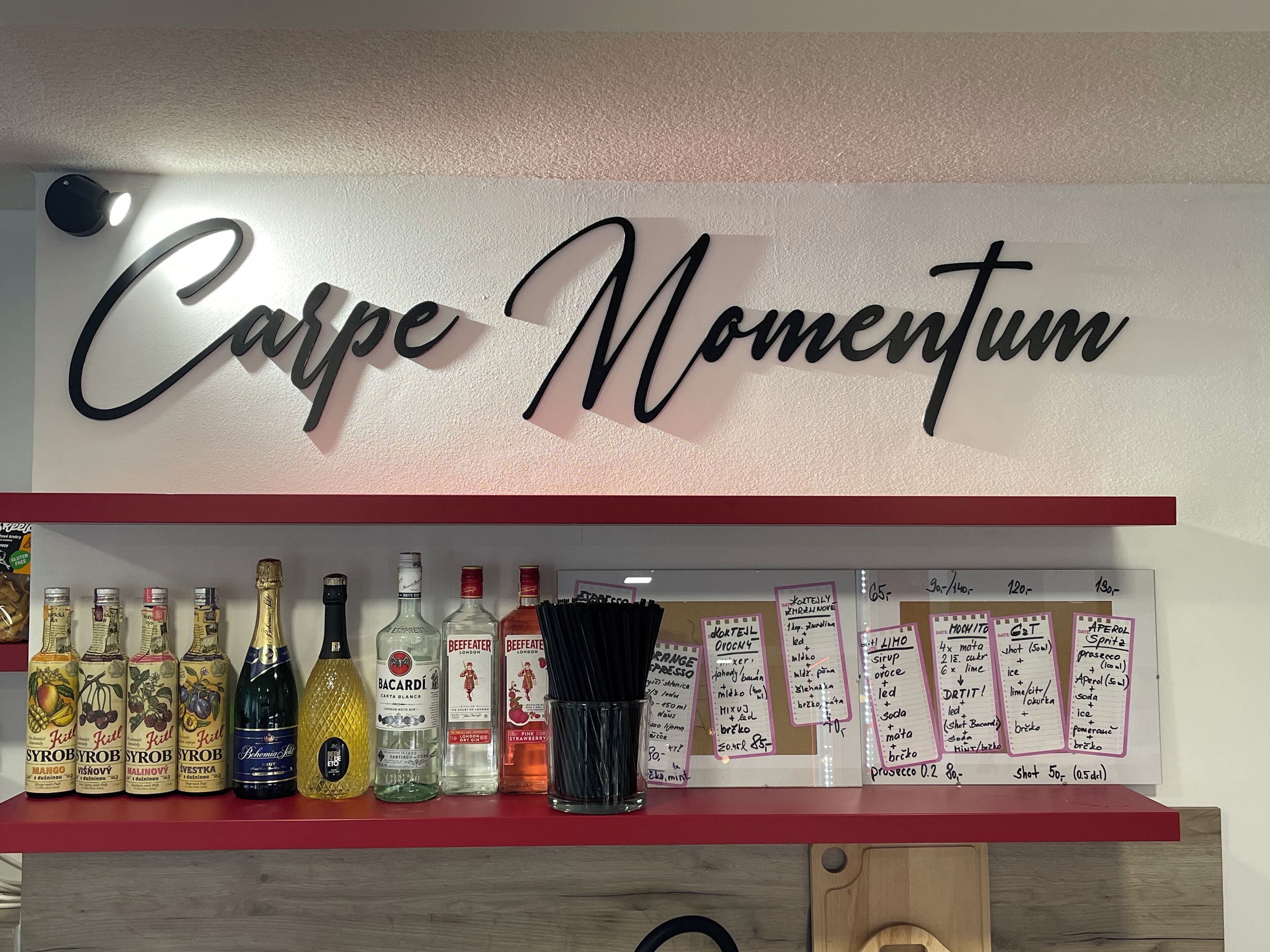 Zona de bar amb un cartell que diu "Carpe Momentum" i prestatges amb diverses ampolles i ingredients per a còctels.