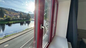 A room with a large window overlooking a river and a park with autumn trees. - 1-bedroom apartment with river view - Greenway Jizera trail, Bohemian Paradise (Železny Brod)