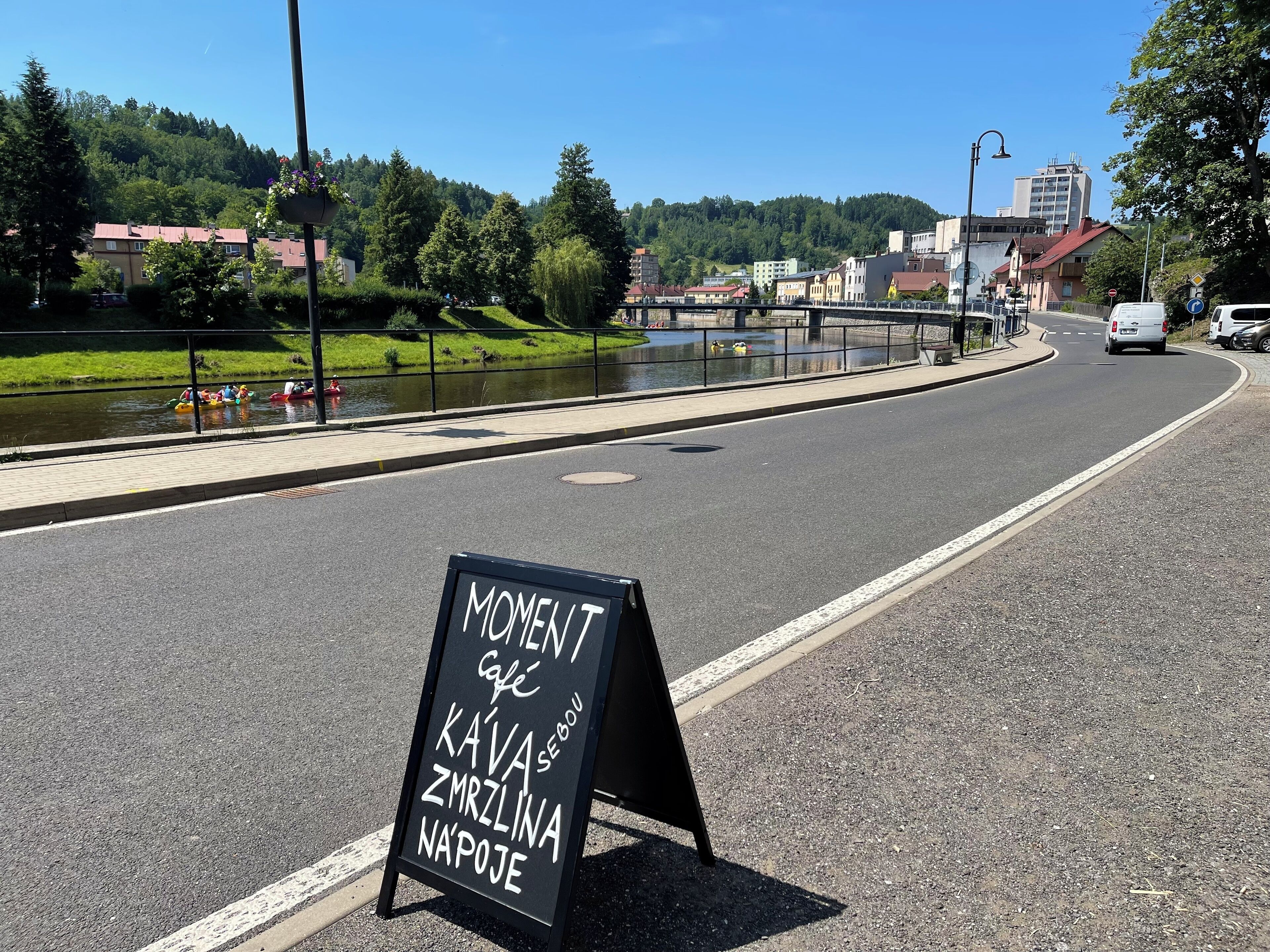A signboard with text in a non-English language, a road with a pedestrian path alongside a river, and buildings in the background.