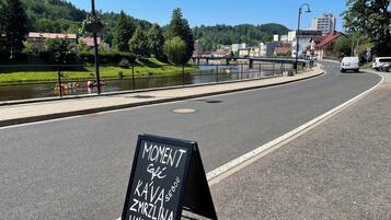 A signboard with text in a non-English language, a road with a pedestrian path alongside a river, and buildings in the background.