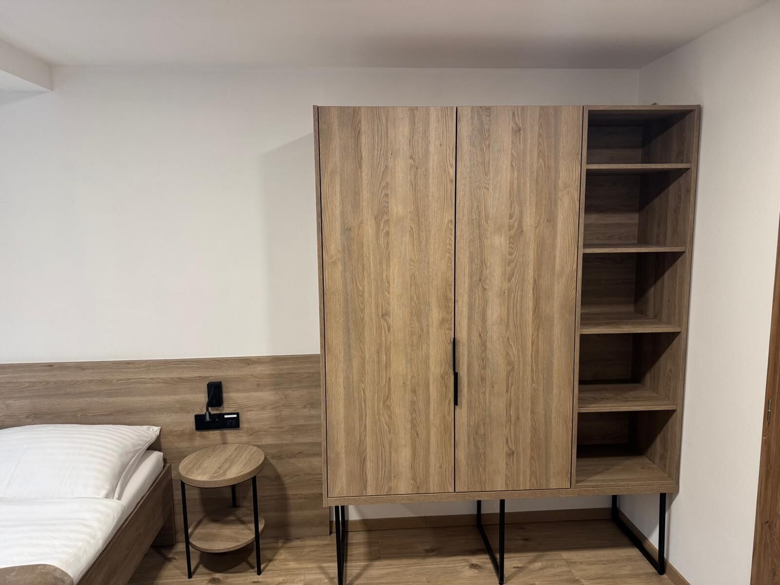 Une chambre à coucher avec un lit, une petite table ronde, une armoire en bois dotée de plusieurs étagères et un objet noir accroché au mur.