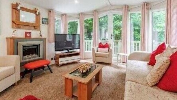 A living room with a fireplace, television, and a view of greenery outside. - Deers Leap - Kelling Heath Holiday Park - 22250 (Holt)
