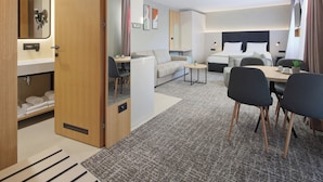 A modern hotel room with a wooden wardrobe, a sofa, a dining table with chairs, and a small kitchen area.