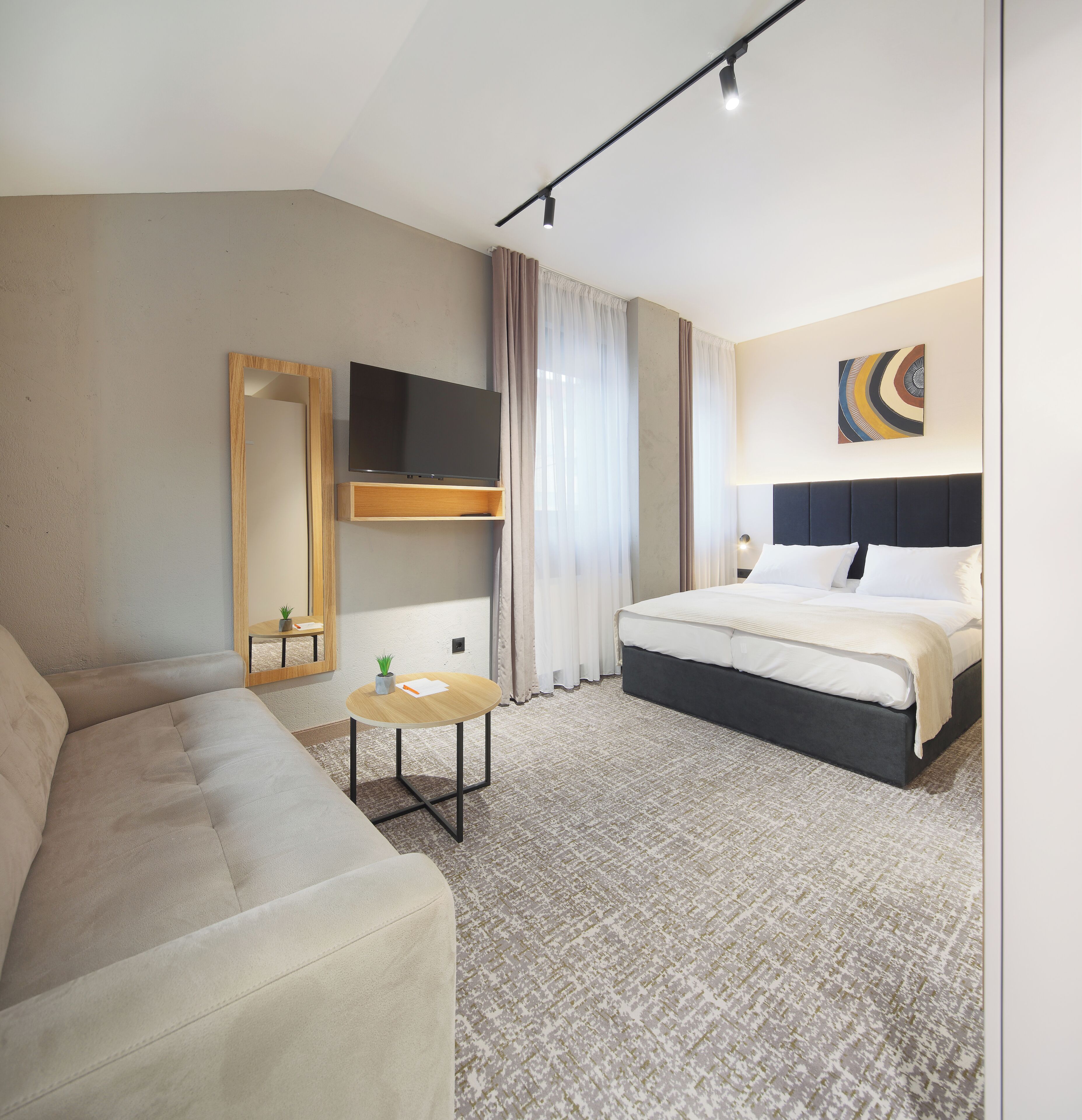 A modern hotel room with a bed, a sofa, a small table, a TV, and a mirror.