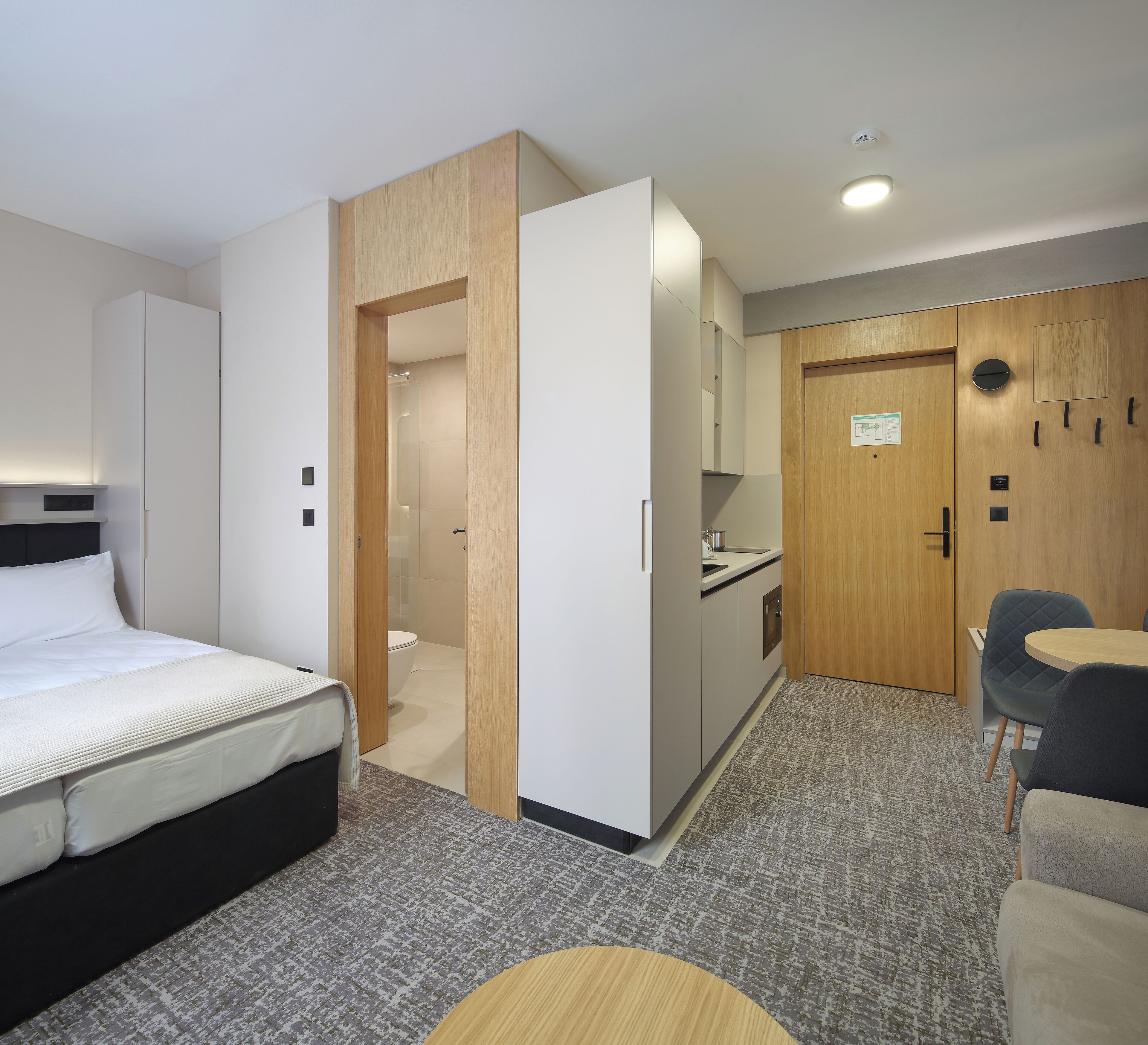 A hotel room with a bed, a small desk, a chair, a kitchenette, and a bathroom.