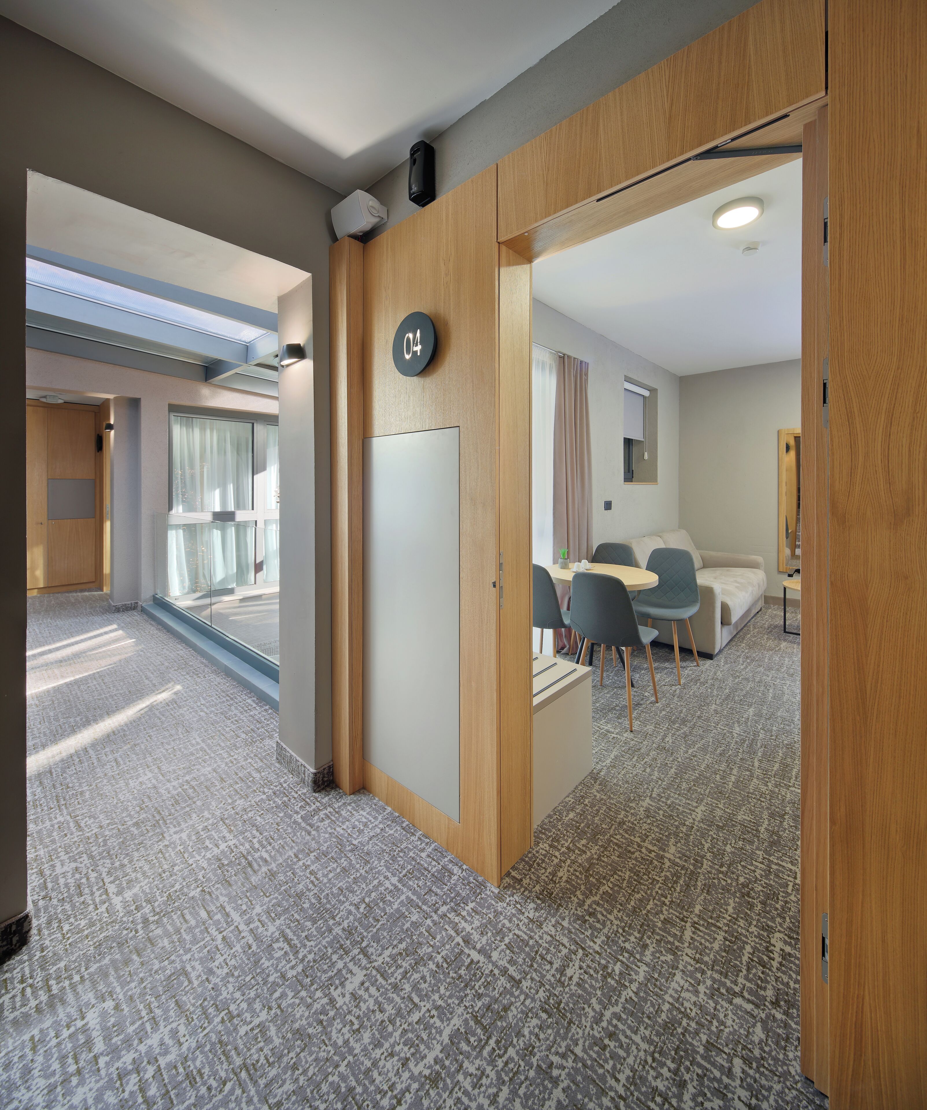 A modern hotel room with a large window, a wooden door with a number sign, a small table with chairs, and a bed.
