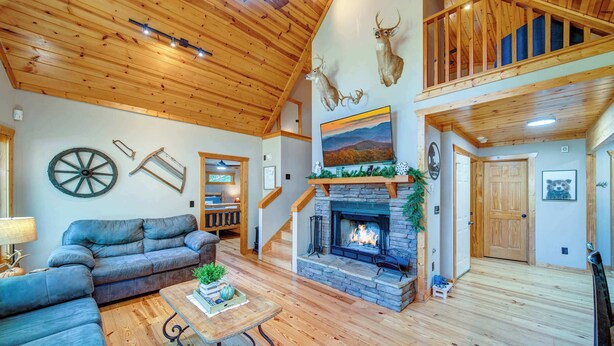 A spacious living room with a fireplace, wooden ceiling, and a loft area with deer antlers on the wall.