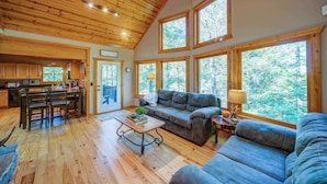 A spacious living area with a wooden ceiling, large windows, a sofa, a coffee table, and a dining area with chairs.
