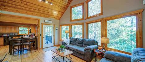 A spacious living area with a wooden ceiling, large windows, a sofa, a coffee table, and a dining area with chairs.