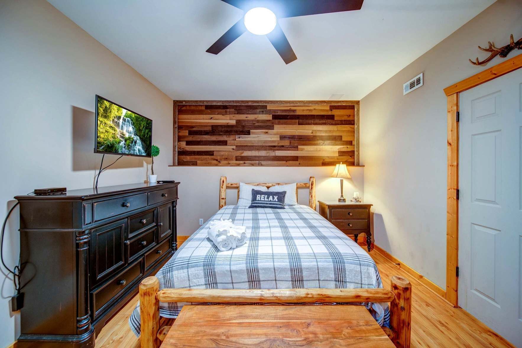 A bedroom with a wooden headboard, a bed with a plaid bedspread, a television on the wall, a dresser, and a ceiling fan.