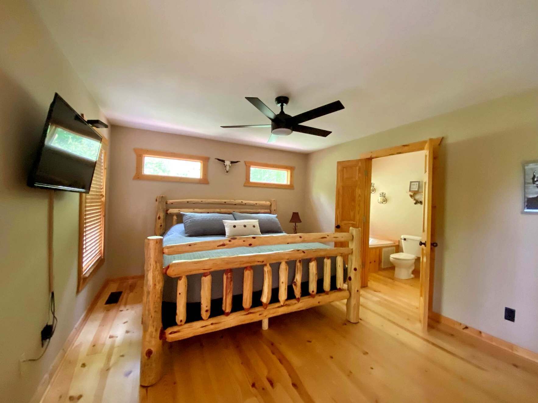 A wooden bed with a headboard, a ceiling fan, a television mounted on the wall, and a bathroom visible through an open door.
