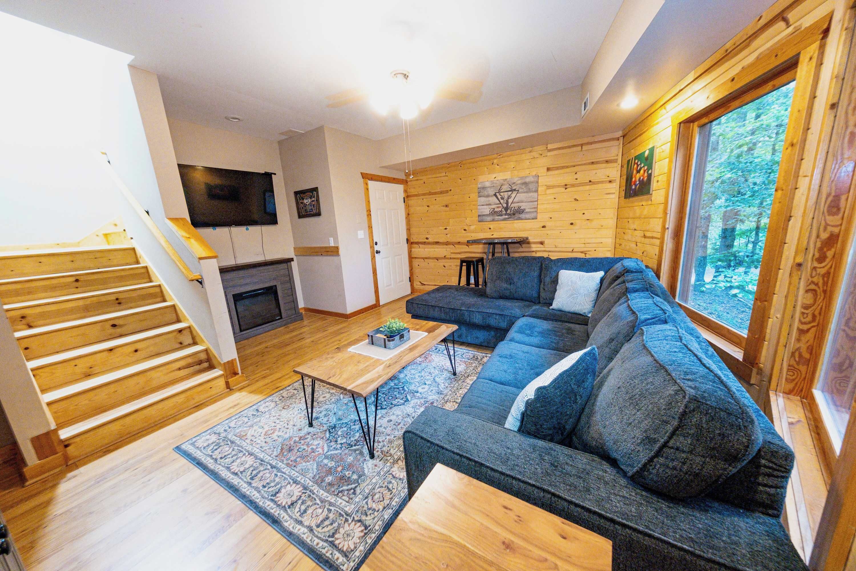 Een moderne woonkamer met een houten trap, een hoekbank, een open haard en een groot raam met uitzicht op de omgeving.