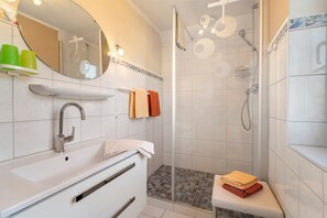 Ein modernes Badezimmer mit Waschbecken, Spiegel, Dusche und einer kleinen Sitzbank mit Handtüchern.