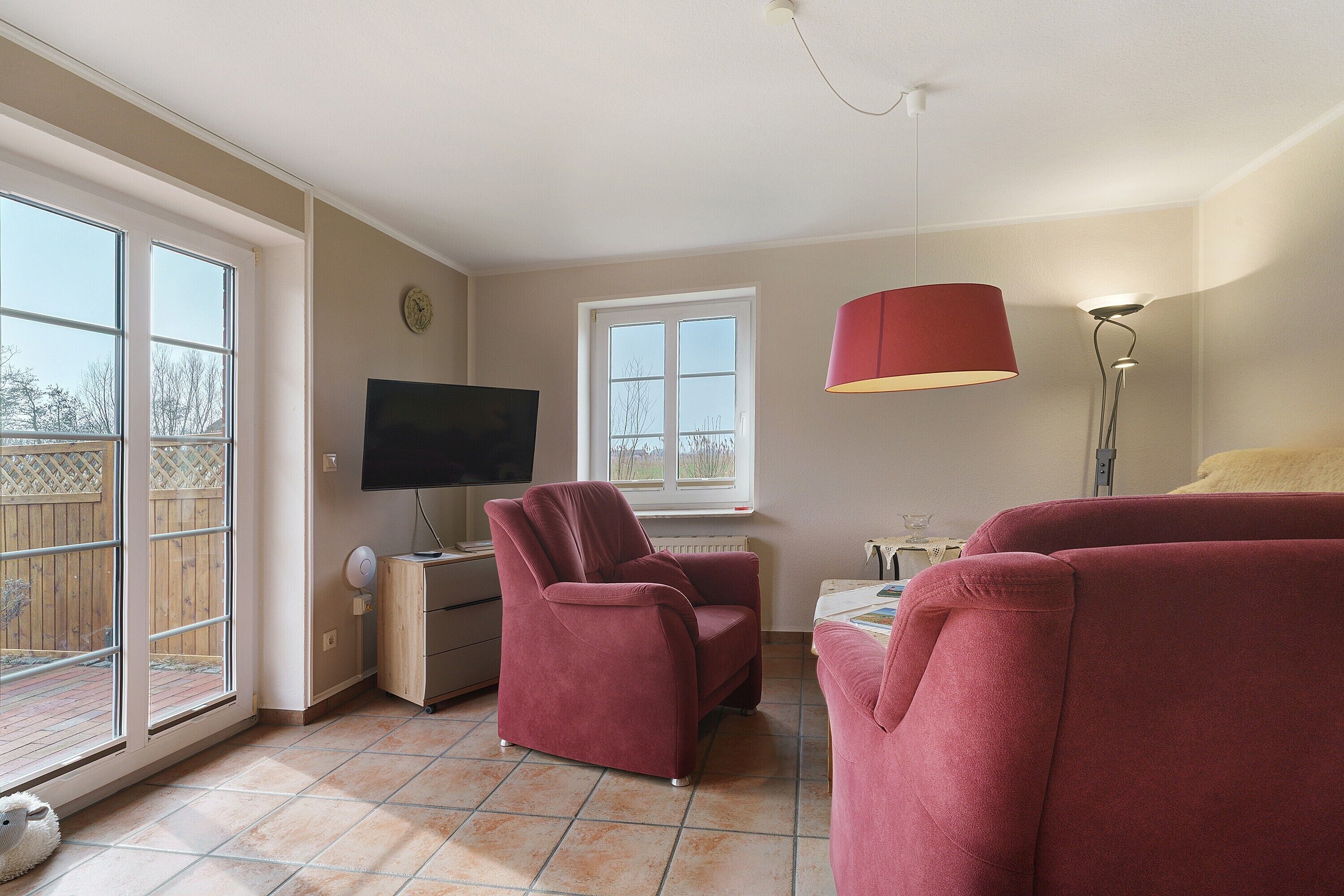 A living room with a TV, a red chair, a red sofa, a lamp, and a window with a view of the outside.