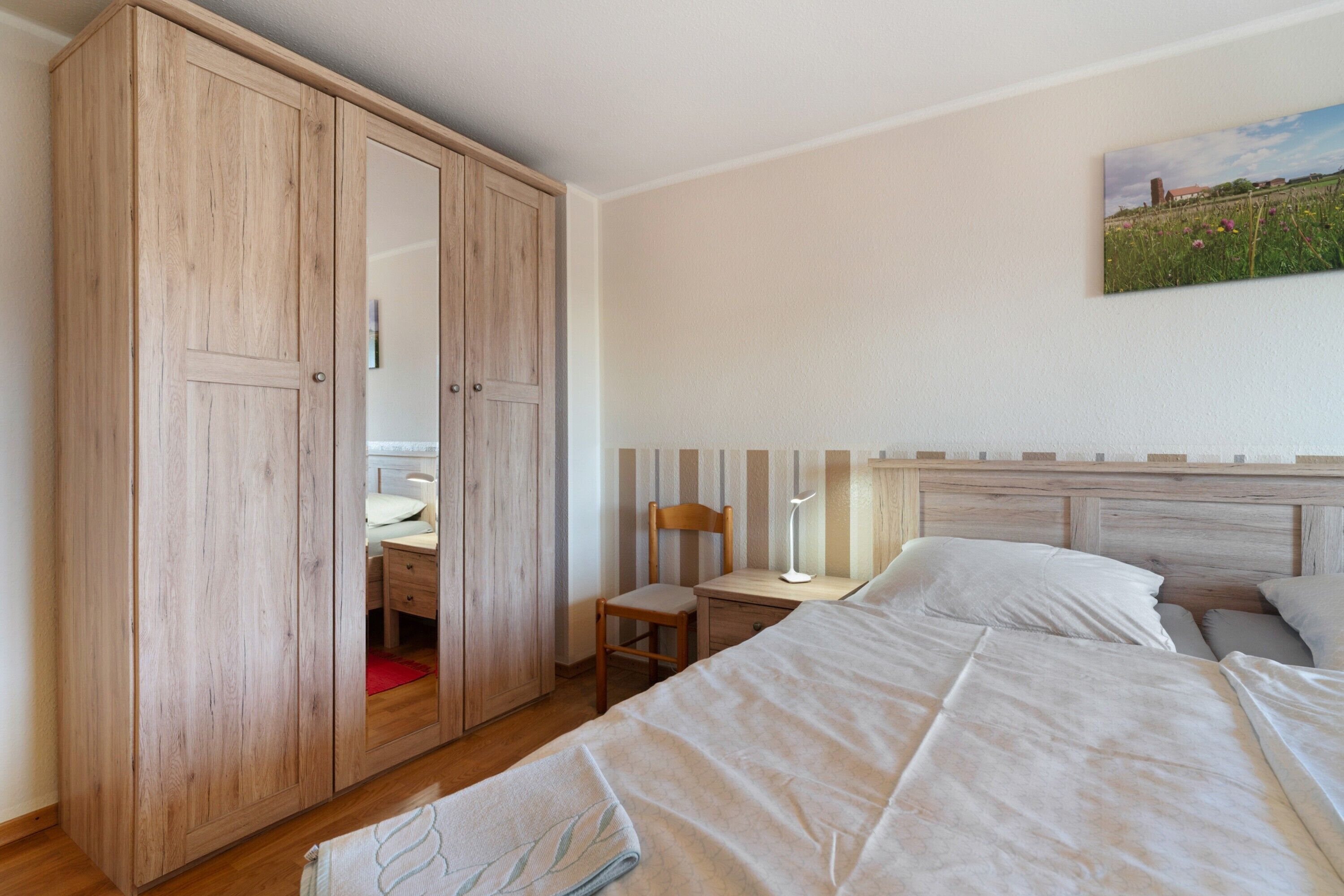 A bedroom with a wooden wardrobe, a desk, a chair, a bed with white bedding, and a painting on the wall.