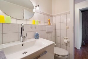 A bathroom with a white sink, mirror, and toilet. There are toiletries on the sink and a towel rack. - Apartment 'Fewo Seegarden' with Shared Garden and Wi-Fi (Pellworm)