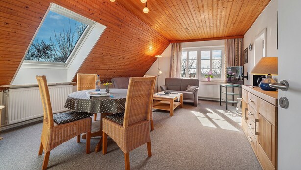 Ein gemütliches Zimmer mit Holzbalkendecke, einem Esstisch mit Stühlen, einer Couch und einem kleinen Tisch mit Lampe.