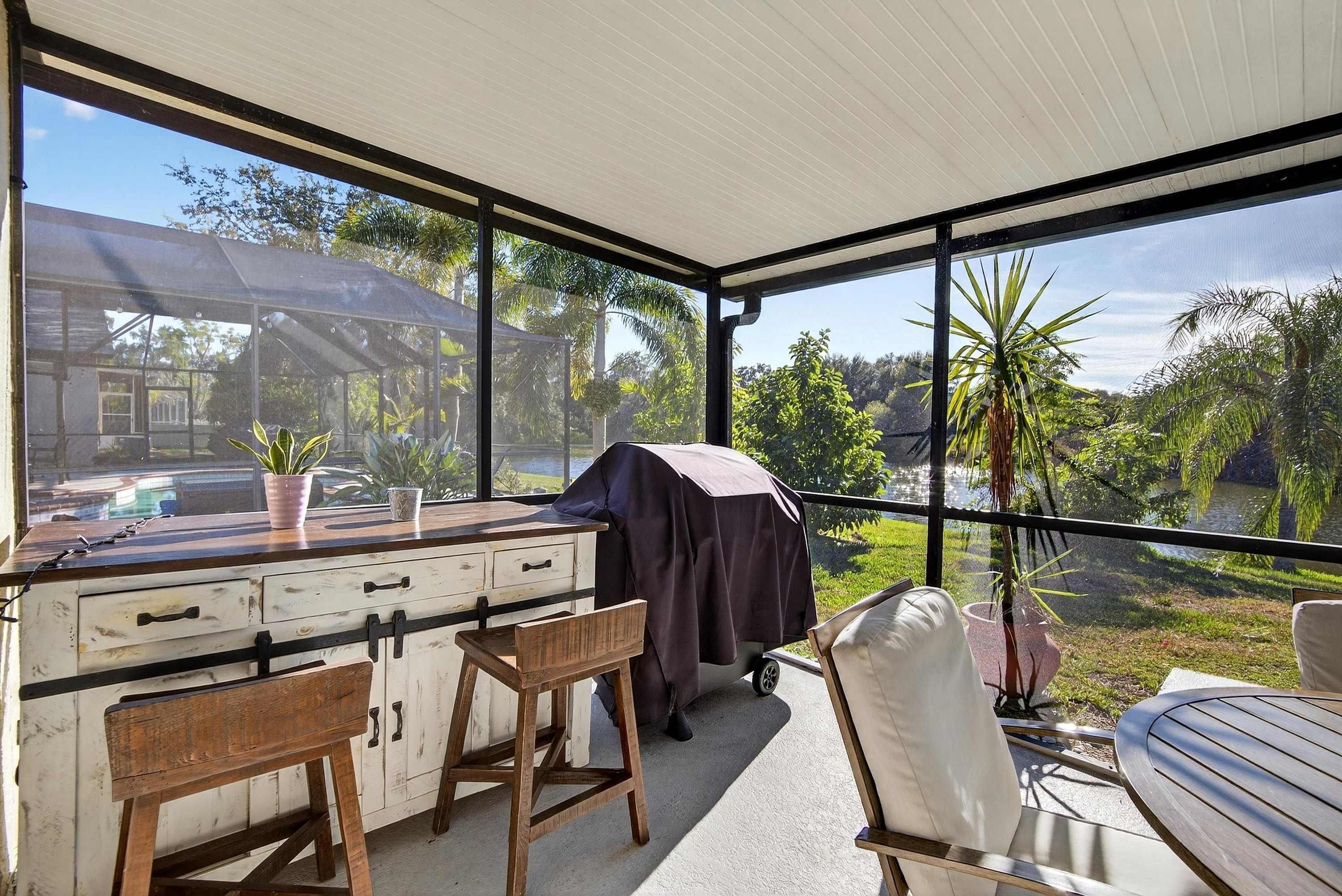 A covered outdoor area with a dining table, chairs, a grill, and a view of a house and greenery.
