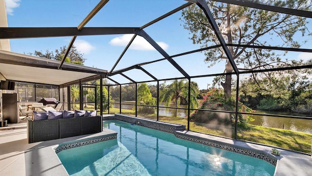 An indoor swimming pool with a glass ceiling, a seating area with cushions, and a view of a lake and trees. - Lakefront 4-bedroom Home With Pool (Brandon)