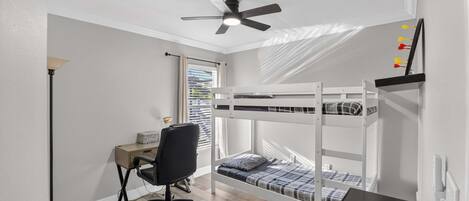 A bedroom with a bunk bed, a desk with a chair, a ceiling fan, and a window with blinds.