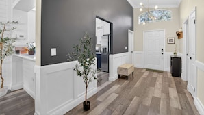 A modern hallway with a dark wall, white trim, and a wooden floor. There is a potted plant, a bench, and a door leading to a kitchen area. - Lakefront 4-bedroom Home With Pool (Brandon)