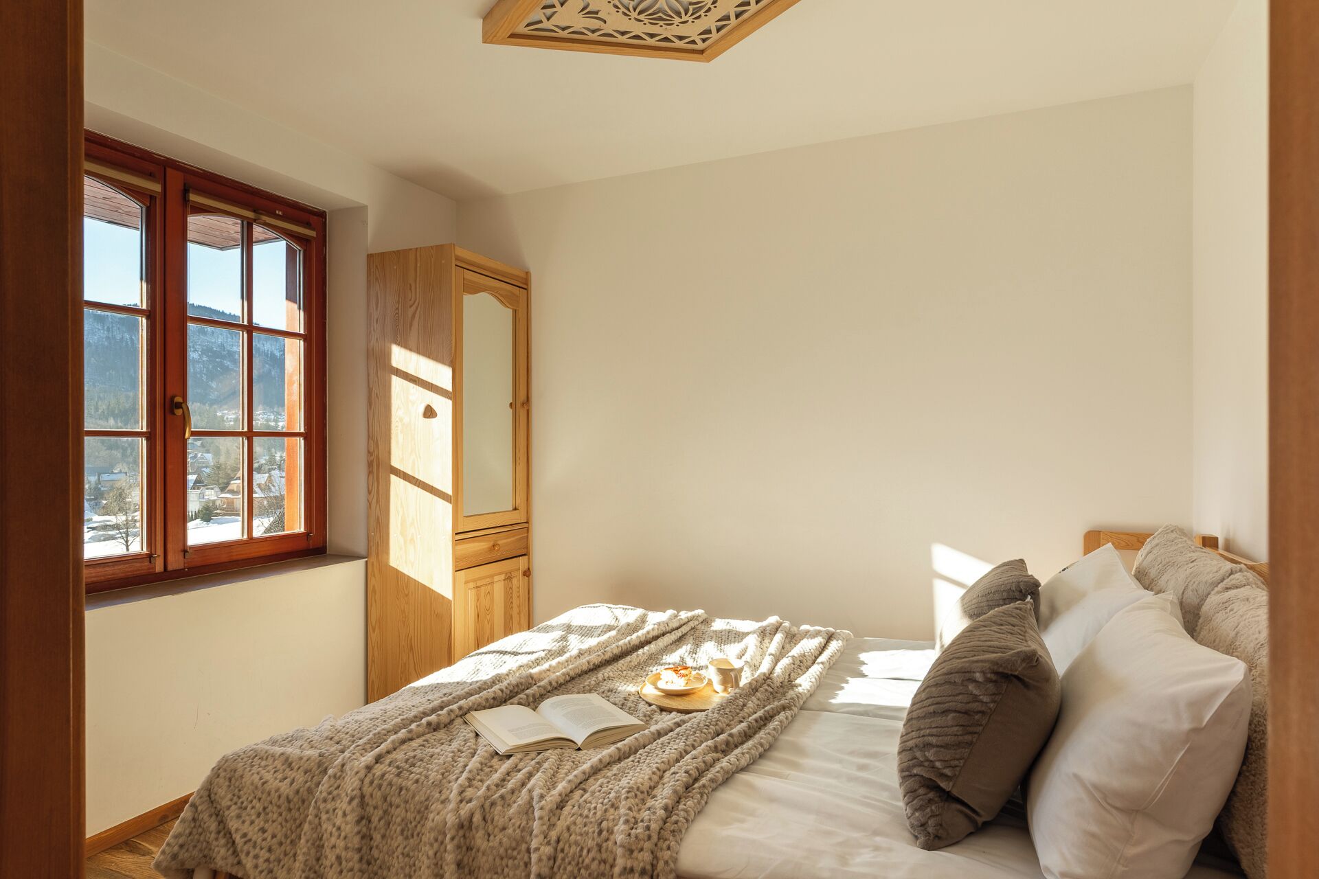 Une chambre avec une grande fenêtre, une armoire en bois, un lit avec une couverture et des oreillers, et un livre posé sur le lit.