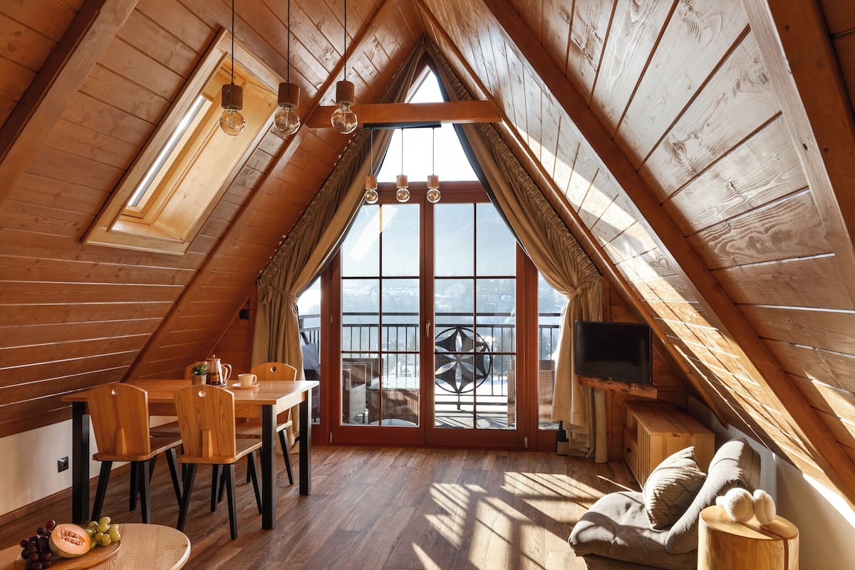 Une pièce mansardée chaleureuse avec des poutres en bois, une table à manger avec des chaises, un canapé et une grande fenêtre donnant sur un balcon.