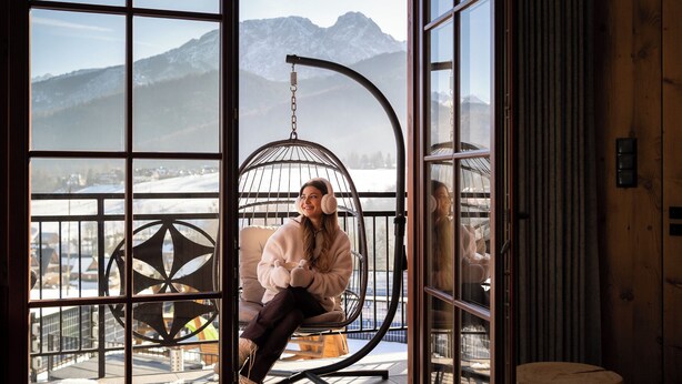 A person sitting on a swing chair by a window overlooking a snowy landscape with mountains. - Karpielówka Suites Parking & Mountain View by Downtown Apartments (Kościelisko)