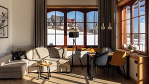A cozy living room with a sofa, a small table, and a desk with chairs. Large windows offer a view of a snowy landscape and a building. - Karpielówka Suites Parking & Mountain View by Downtown Apartments (Kościelisko)