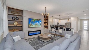 A modern living room with a grey sofa set, a fireplace, a TV mounted on the wall, and a kitchen area in the background. - 9 Bedroom With Everything For Happiness! 9 Home (Kissimmee)