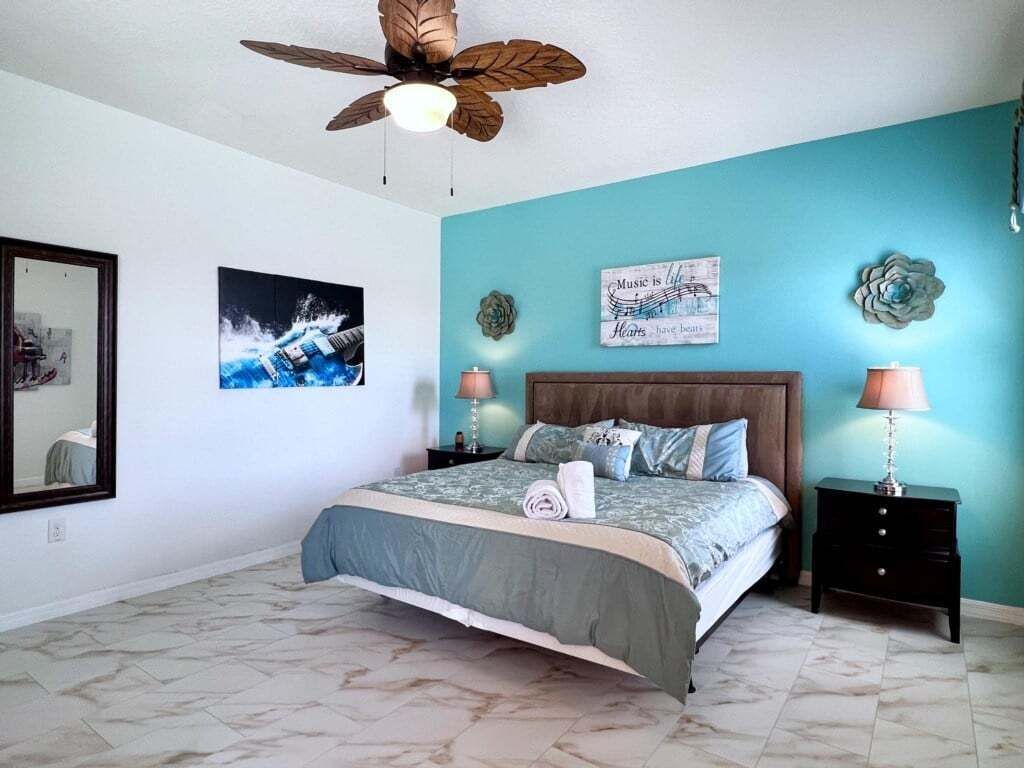 A bedroom with a bed, a ceiling fan, a wall with a poster, and a nightstand with a lamp.