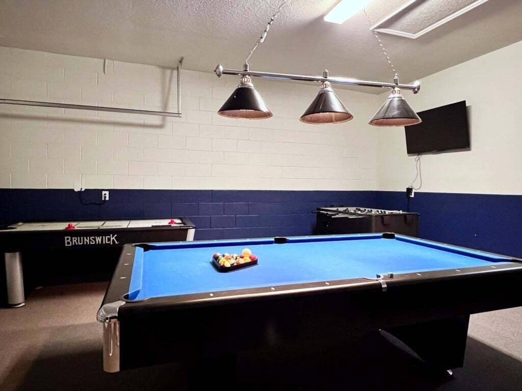 A pool table with a blue felt surface, three hanging lamps, and a television mounted on the wall.