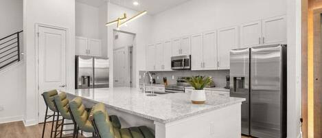 A modern kitchen with white cabinetry, stainless steel appliances, a central island with bar stools, and a hanging light fixture.