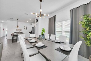 Un espace repas moderne avec une longue table dressée pour six personnes, des chaises blanches, un lustre et une cuisine en arrière-plan.