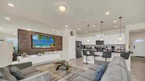 A modern living room with a large flat-screen TV, grey sofas, a coffee table, and a kitchen in the background. - Lake View Serene & Peaceful Pool Home! 5 Bedroom Townhouse (Kissimmee)