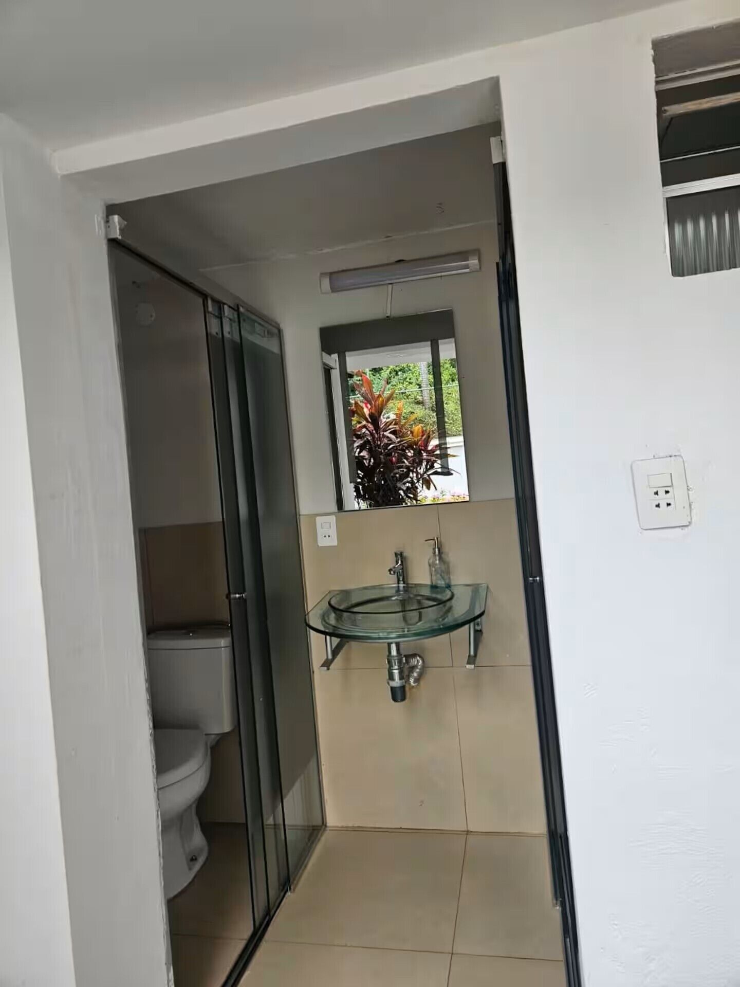 Ein kleines Badezimmer mit einem Waschbecken aus Glas, einer Toilette und einem Fenster mit Blick ins Grüne.
