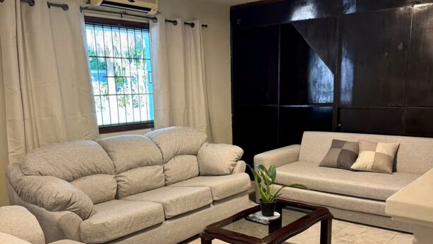 Una sala de estar con un sofá seccional color beige, un sofá más pequeño, una mesa de centro de vidrio con una planta y una ventana con cortinas.