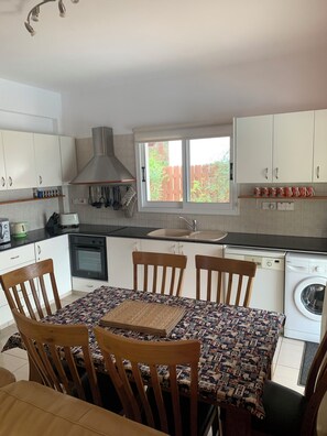 A modern kitchen with white cabinets, a built-in oven, a stove, a sink, and a washing machine. There is a dining table with chairs and a window with a view of a wooden fence. - Villa Eldora Lysos With Beautiful pool (Lysos)