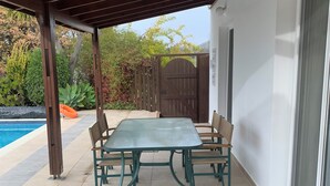 A covered patio with a table and chairs, a pool, and a gate leading to a garden.