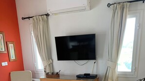 A living room with a beige leather sofa, a wooden cabinet, a glass coffee table, a television, and a window with curtains.