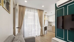 Un soggiorno moderno con un divano, una TV a schermo piatto montata su una parete color verde acqua, e una cucina sullo sfondo.