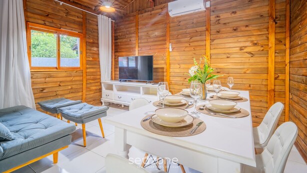 A wooden cabin interior with a dining table set for four, a sofa, a TV, and an air conditioning unit. - Wooden Retreat Near Lagoa da Conceicao Knu0170 (Florianópolis)