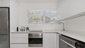 Ett modernt kök med vita köksskåp, ett kylskåp i rostfritt stål, en inbyggd ugn, en diskmaskin och ett fönster med persienner.