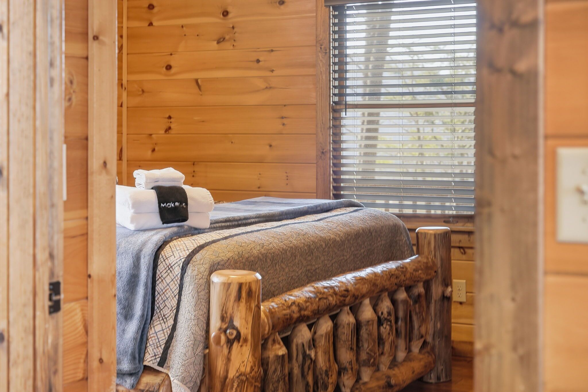 A wooden cabin room with a bed, a window with blinds, and a wooden headboard.