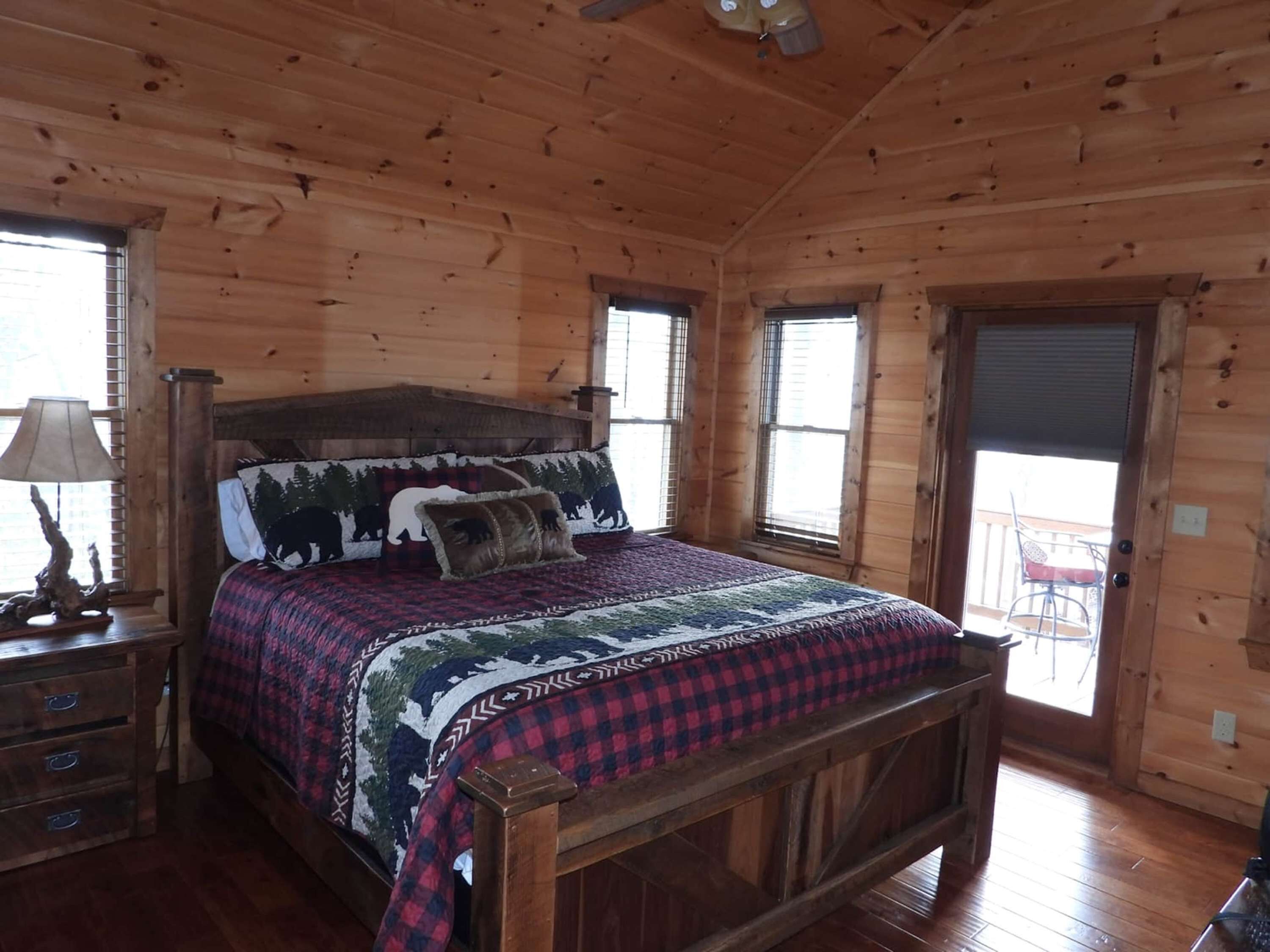Un dormitorio de cabaña de madera con una cama, una mesita de noche, una lámpara y una ventana con persianas.