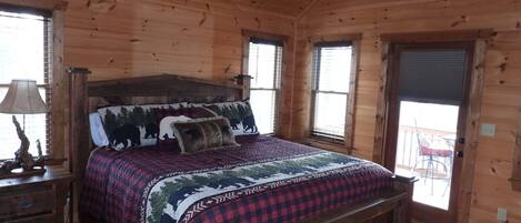 Un dormitorio de cabaña de madera con una cama, una mesita de noche, una lámpara y una ventana con persianas.